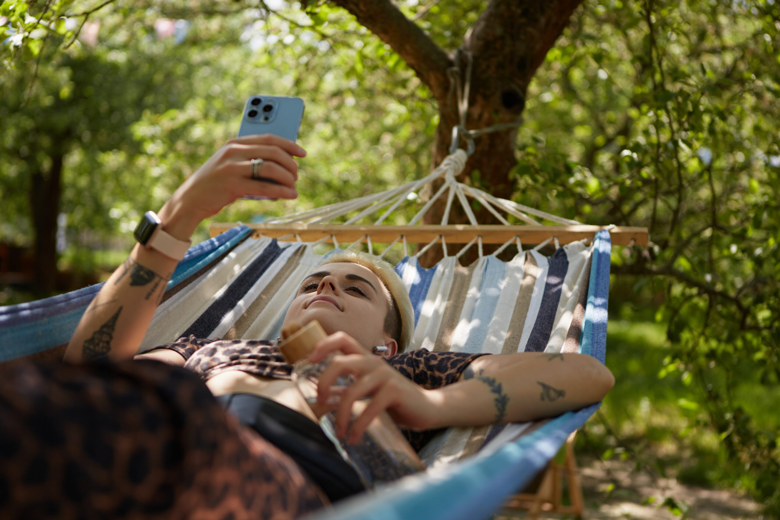 Young diverse woman with short hair lying on a hammock in a park and browsing mobile app on a smartphone. Stylish tattooed female relaxing in a green garden on weekend in summer